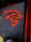 Illuminated Hellcat emblem in red light shining from the open door of a Dodge Charger, casting a sharp reflection on a concrete floor.