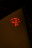 Close-up photo of a red Hellcat logo projection on the ground, illuminated by the door light of a Dodge Challenger SRT Hellcat.